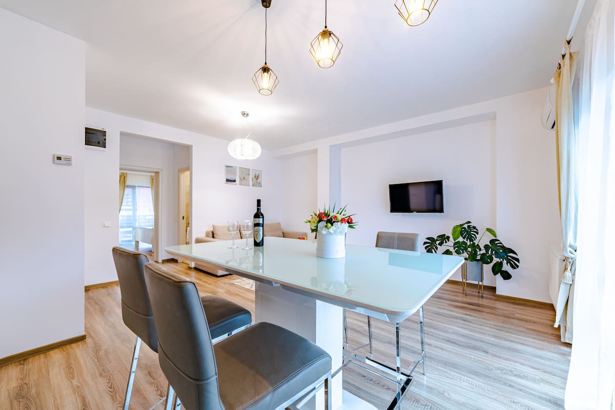 A modern living room with a glass dining table, comfortable seating, a flat-screen TV, and a potted plant.