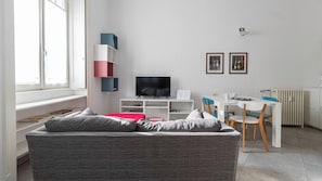 A modern living room with a grey sofa, a flat-screen TV, a dining table with chairs, and a white radiator.