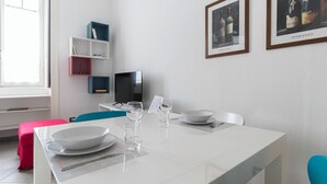 Uma área de jantar moderna com uma mesa branca posta para dois, uma televisão e uma estante de livros.