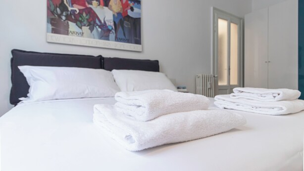 Uma cama bem arrumada com lençóis e toalhas brancas, uma cabeceira preta e um quadro na parede.