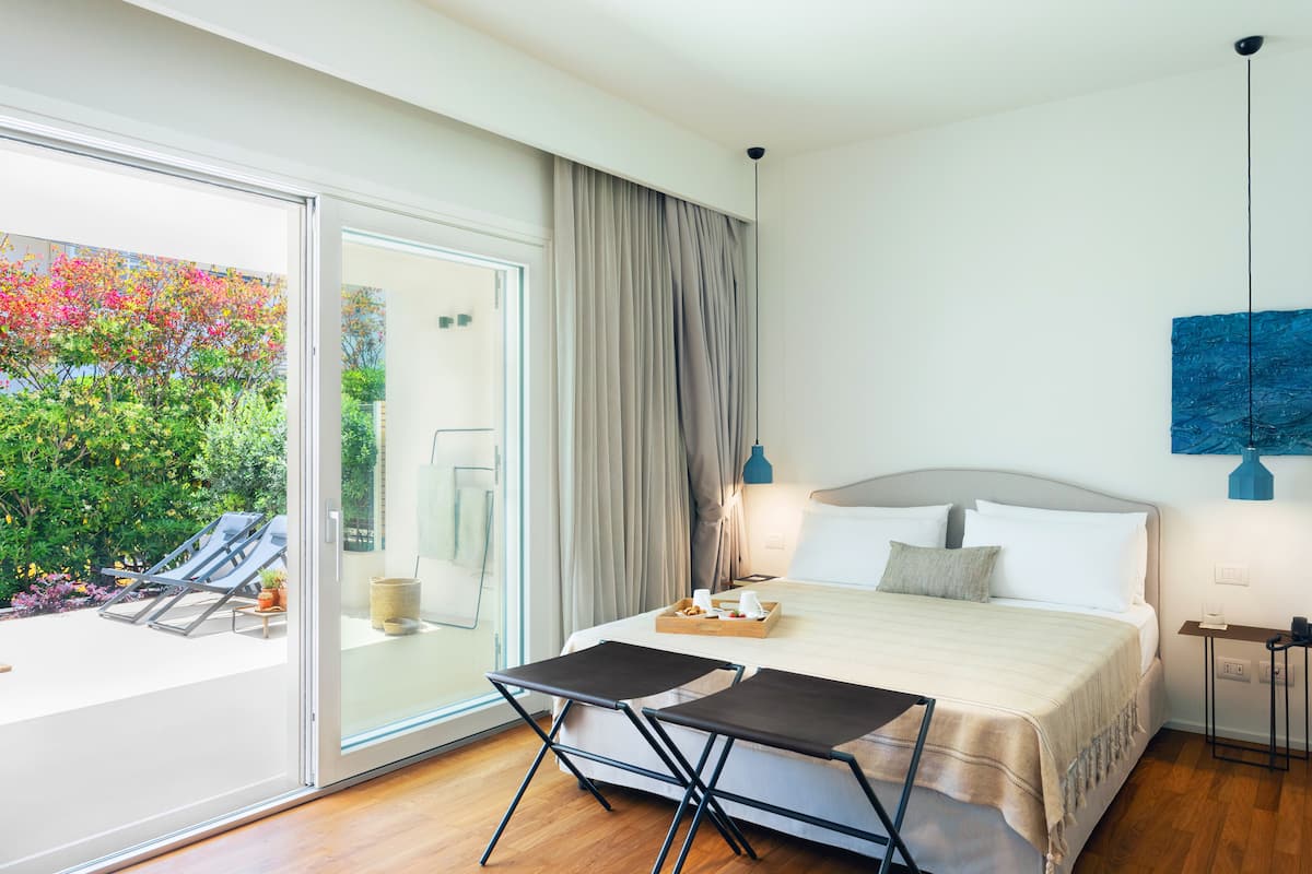 A modern hotel room with a large bed, a dining table, and a view of a garden with colorful plants.