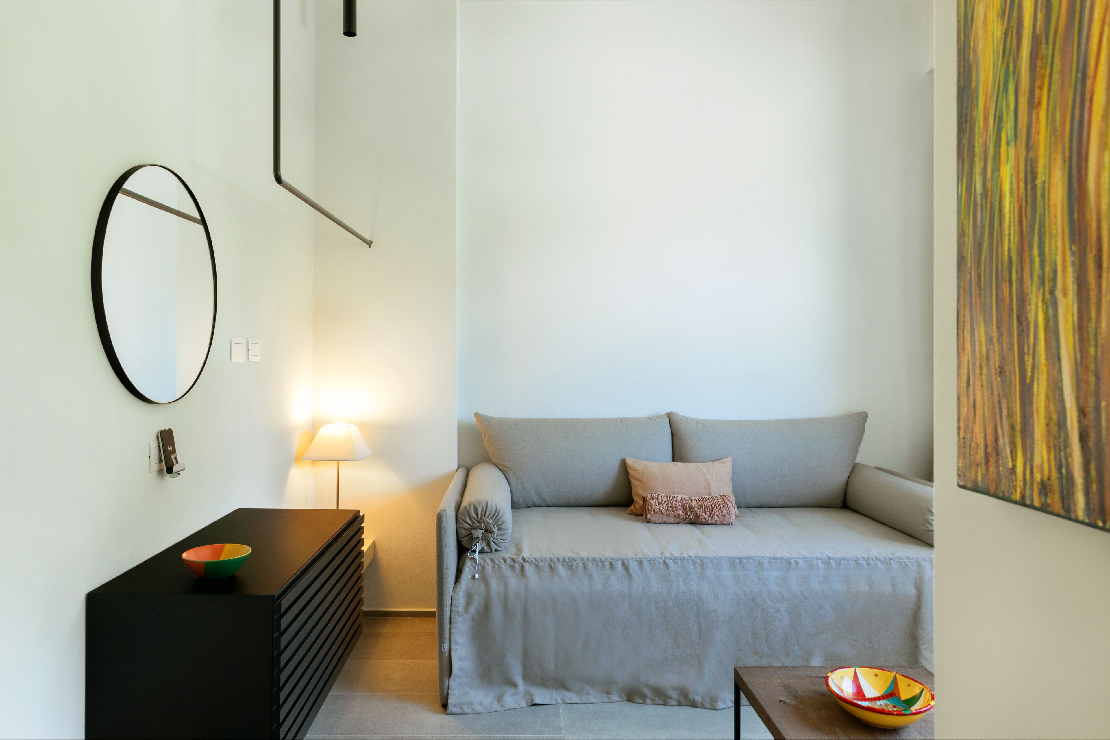 A modern, minimalist bedroom with a sofa, a bedside table, a lamp, and a wall-mounted mirror.