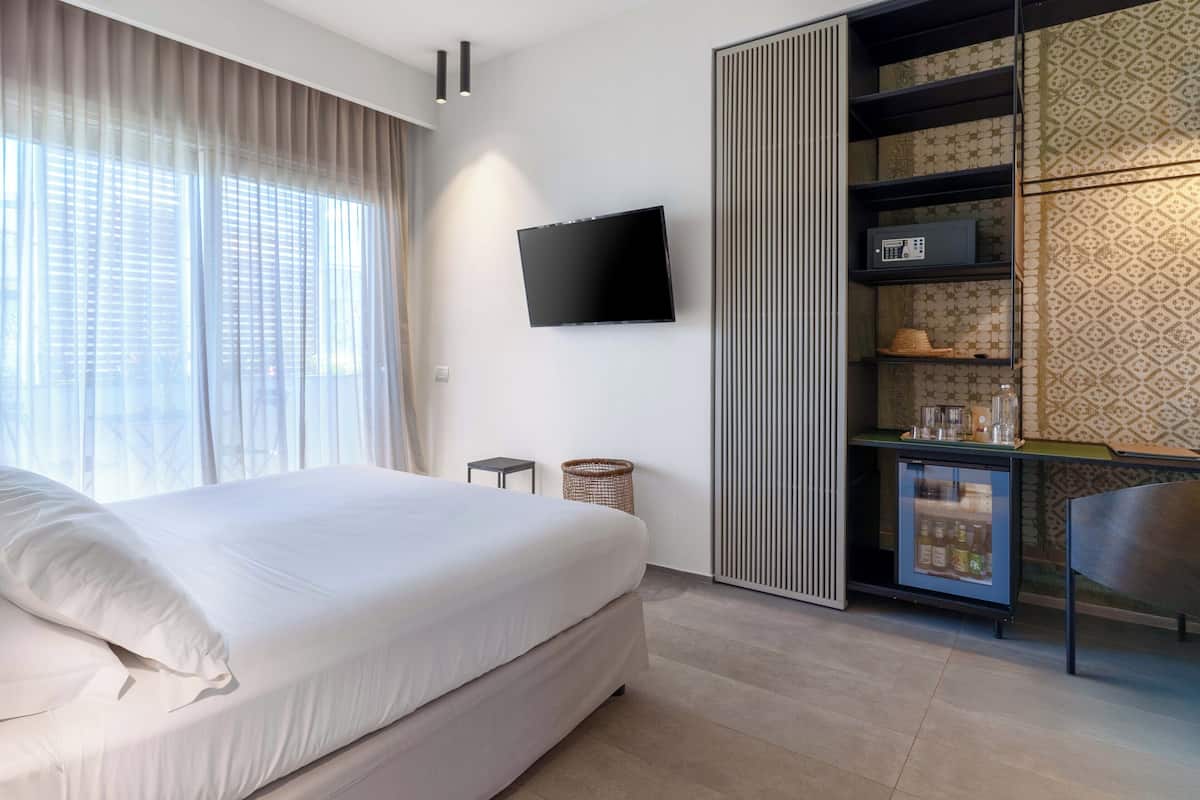 A modern hotel room with a large bed, a flat-screen TV mounted on the wall, a small side table, a woven basket, a minibar, and a patterned wall.