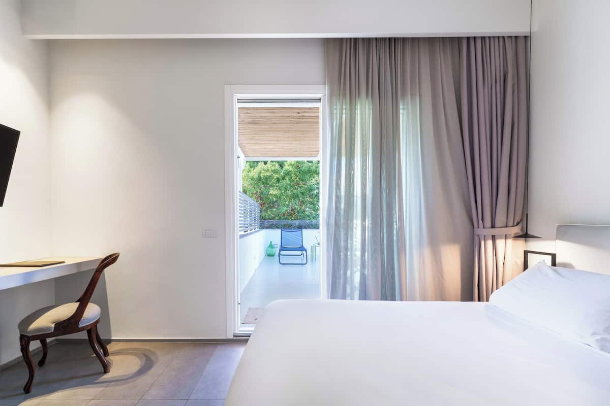 A hotel room with a bed, a desk, a chair, and a view of a balcony with a chair and greenery outside.
