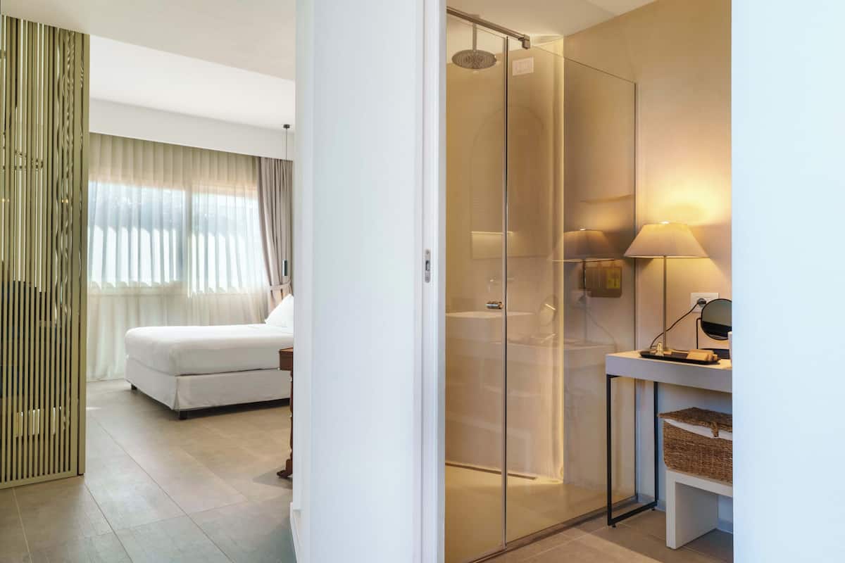 A modern hotel room with a glass shower enclosure, a white bed, and a bedside table with a lamp.