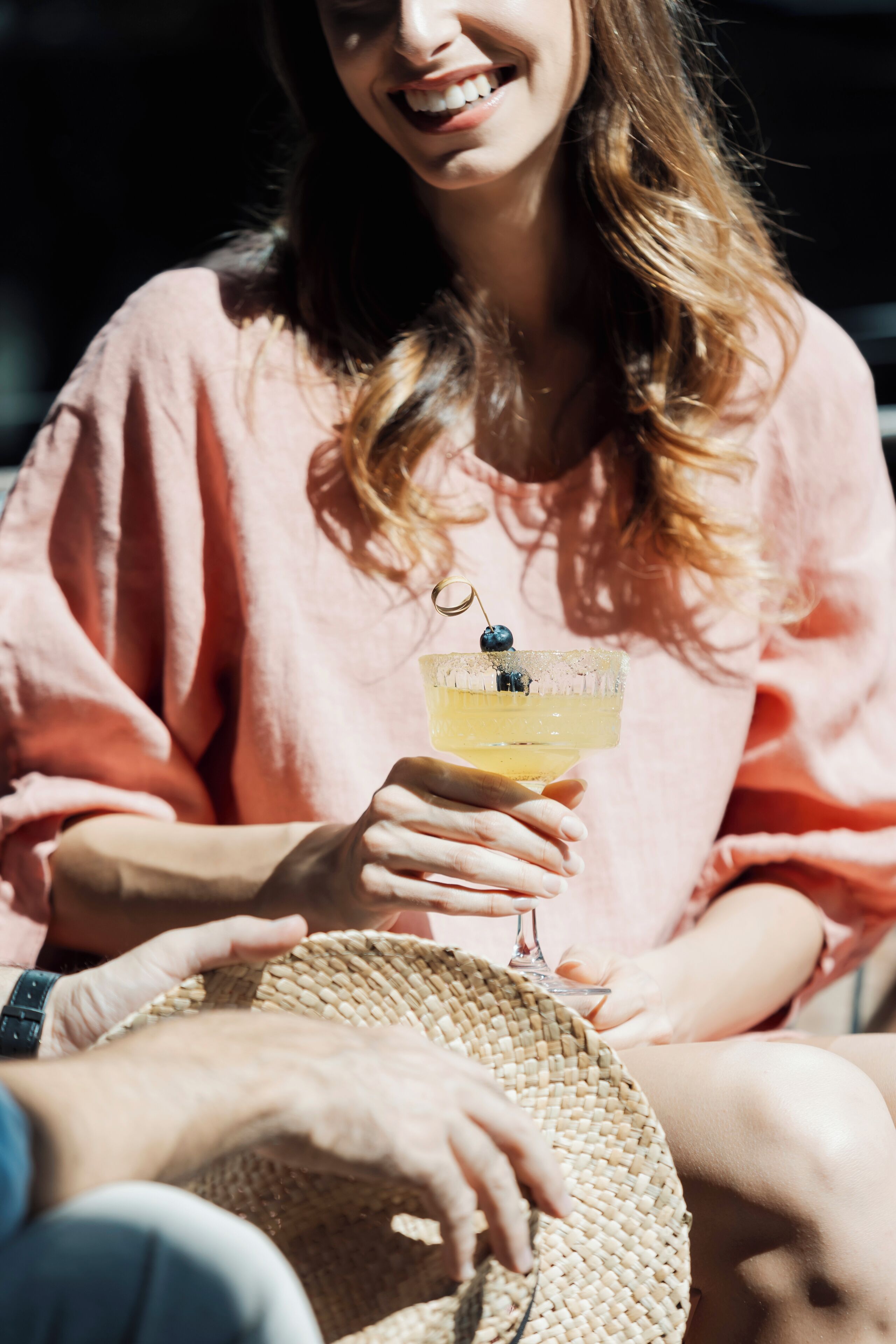 A person holding a drink with a straw and a blueberry garnish, wearing a pink top, and sitting with a straw hat.