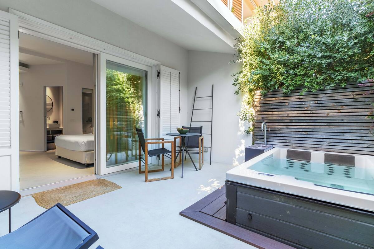 A modern outdoor patio with a hot tub, a dining area with chairs, and a sliding glass door leading to a bedroom.