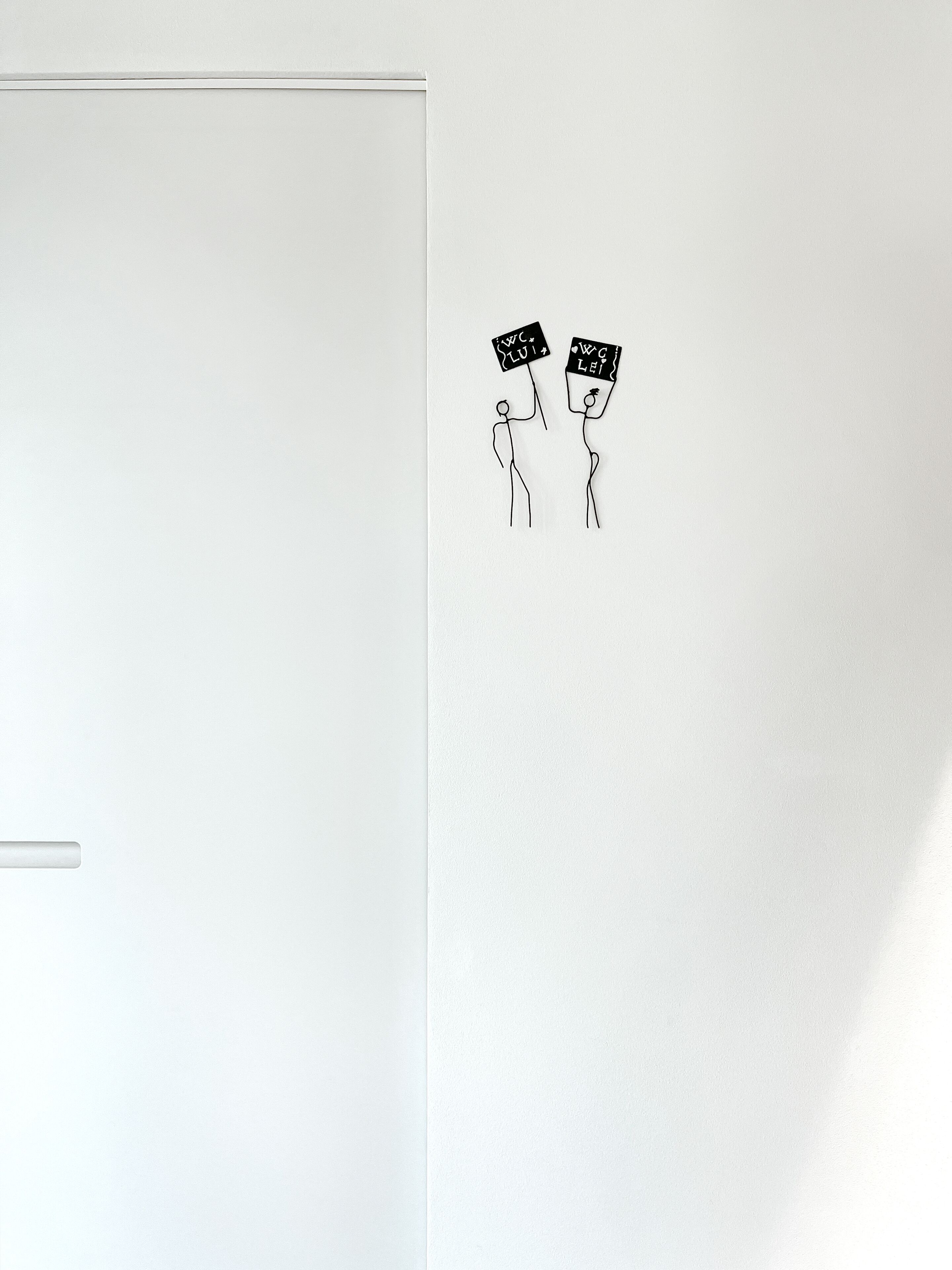 Una pared blanca con dos figuras esquemáticas sosteniendo letreros que dicen "W.C. LUI" y "W.C. LEI".