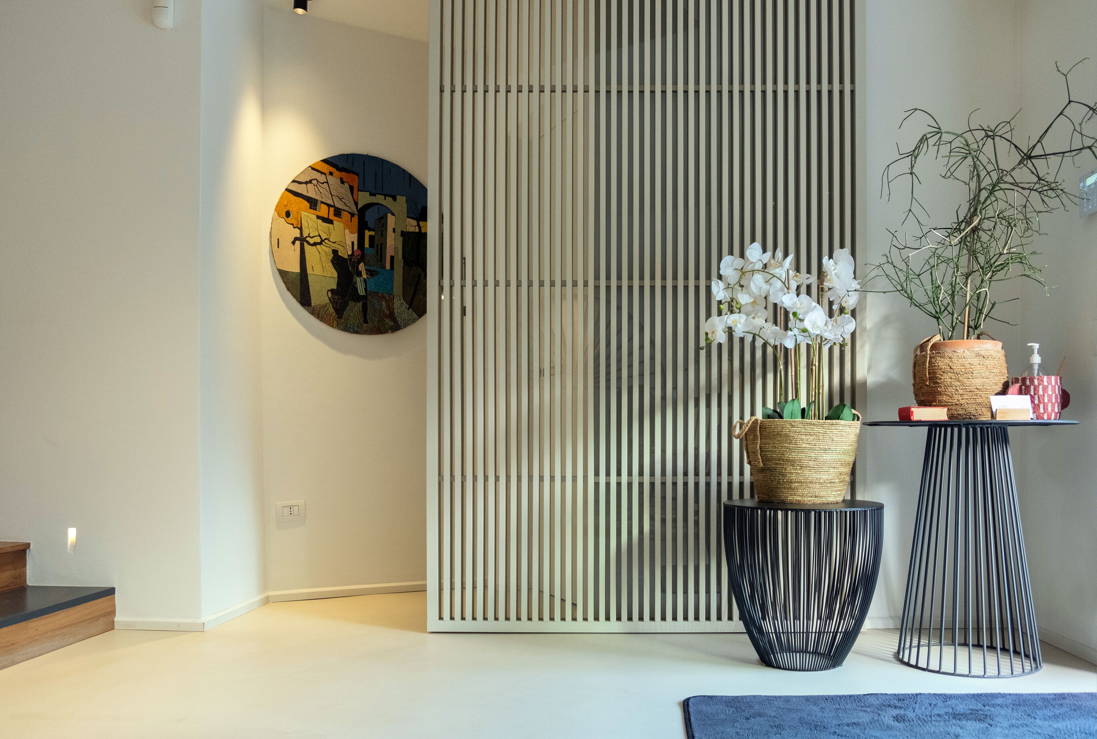 Un interior moderno con diseño minimalista, que incluye una obra de arte circular en la pared, un panel divisor de listones verticales y una pequeña mesa con una planta y una botella.