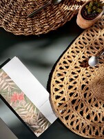 Un cesto intrecciato con un cucchiaio, una pianta in vaso e un cartoncino con motivi a foglie tropicali.