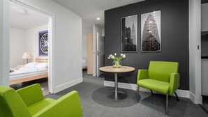 Une chambre d’hôtel avec un lit, un fauteuil vert, une petite table sur laquelle est posé un vase de fleurs, et deux tableaux encadrés accrochés au mur.