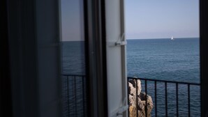 Vista do mar por uma janela com grade de metal, um veleiro ao longe e um afloramento rochoso em primeiro plano.