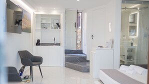 Quarto de hotel moderno com uma cama, área de estar com uma cadeira, pequena cozinha e banheiro com chuveiro de vidro.