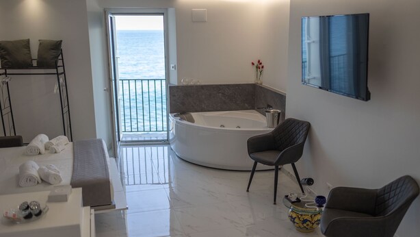 Um banheiro moderno com uma banheira grande, uma área de estar com duas cadeiras, uma mesa pequena com um vaso e vista para o mar por uma porta aberta.