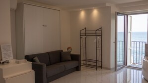 Uma sala de estar moderna com um sofá, uma mesa pequena e uma porta deslizante que dá para uma varanda com vista para o mar.