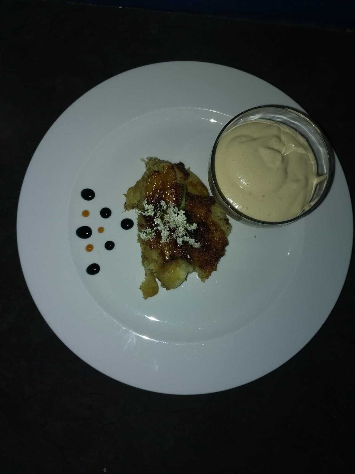 Un plato blanco con un postre, un pequeño recipiente con salsa y algunos puntos negros y amarillos sobre un fondo oscuro.