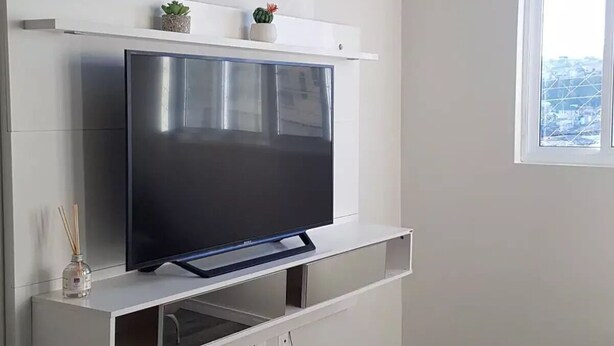 A modern living room with a flat-screen TV mounted on a white wall, a red rug on the floor, and a window with a view of buildings.