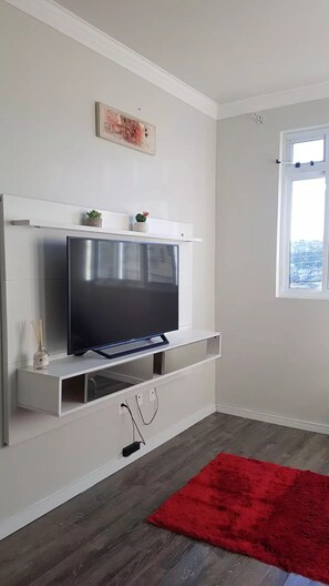 Een moderne woonkamer met een flatscreen televisie aan een witte muur, een rood tapijt op de vloer en een raam met uitzicht op gebouwen.