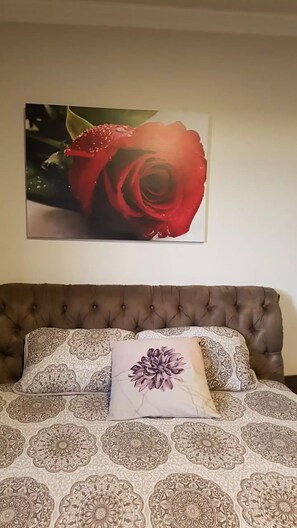 A bedroom with a tufted headboard, patterned bedding, and a large floral painting on the wall. - Apartamento Completo no Centro de Lages (Centro Lages)