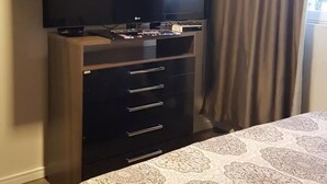 A hotel room with a flat-screen TV mounted on a wooden cabinet, a bed with a patterned bedspread, and a window with curtains.