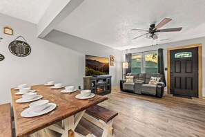 Un comedor con mesa de madera dispuesta para cuatro personas, un sofá, chimenea y puerta que da al exterior.
