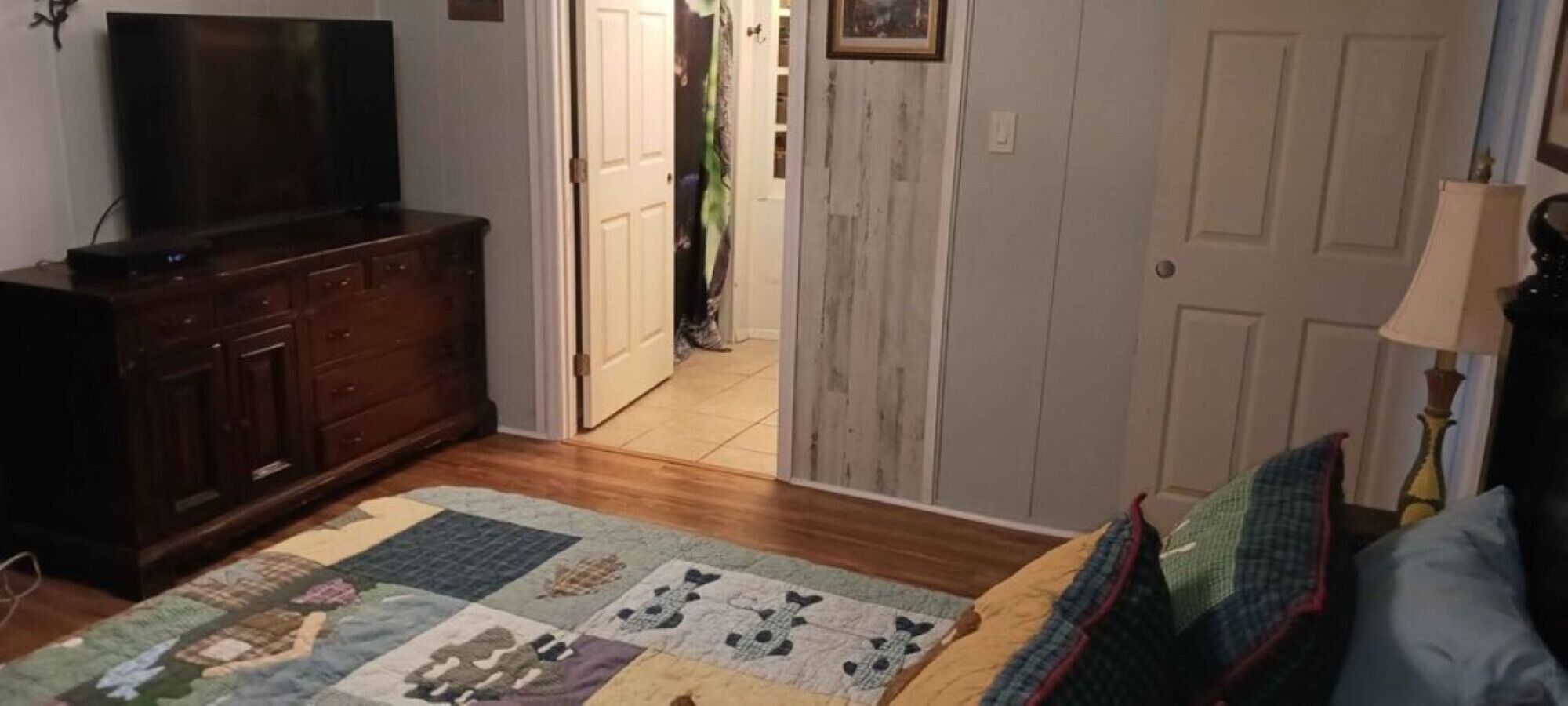 Quarto com cômoda de madeira, televisão, colcha na cama e porta que dá para outro ambiente.