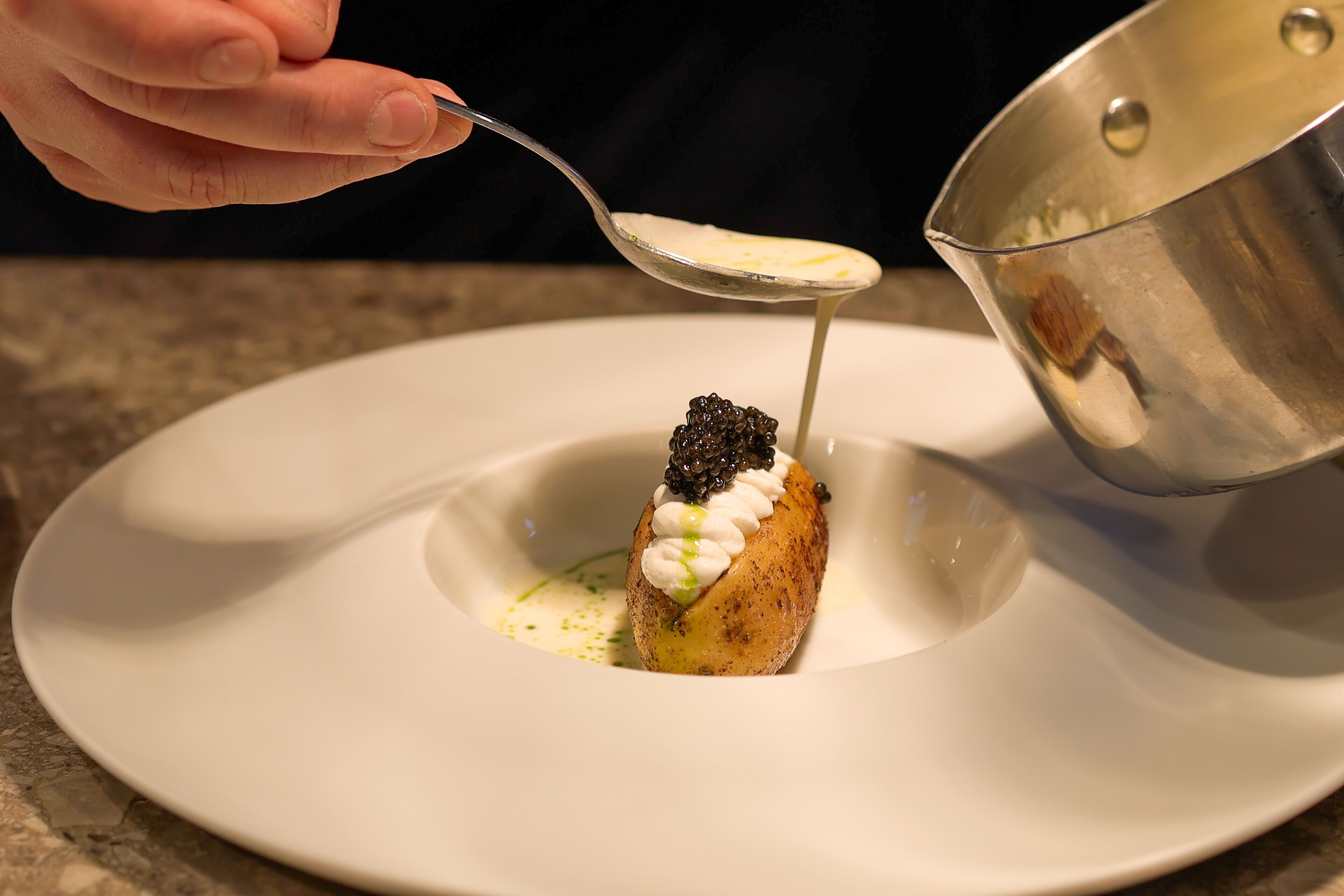 A spoonful of sauce being poured onto a dessert with caviar on a white plate.