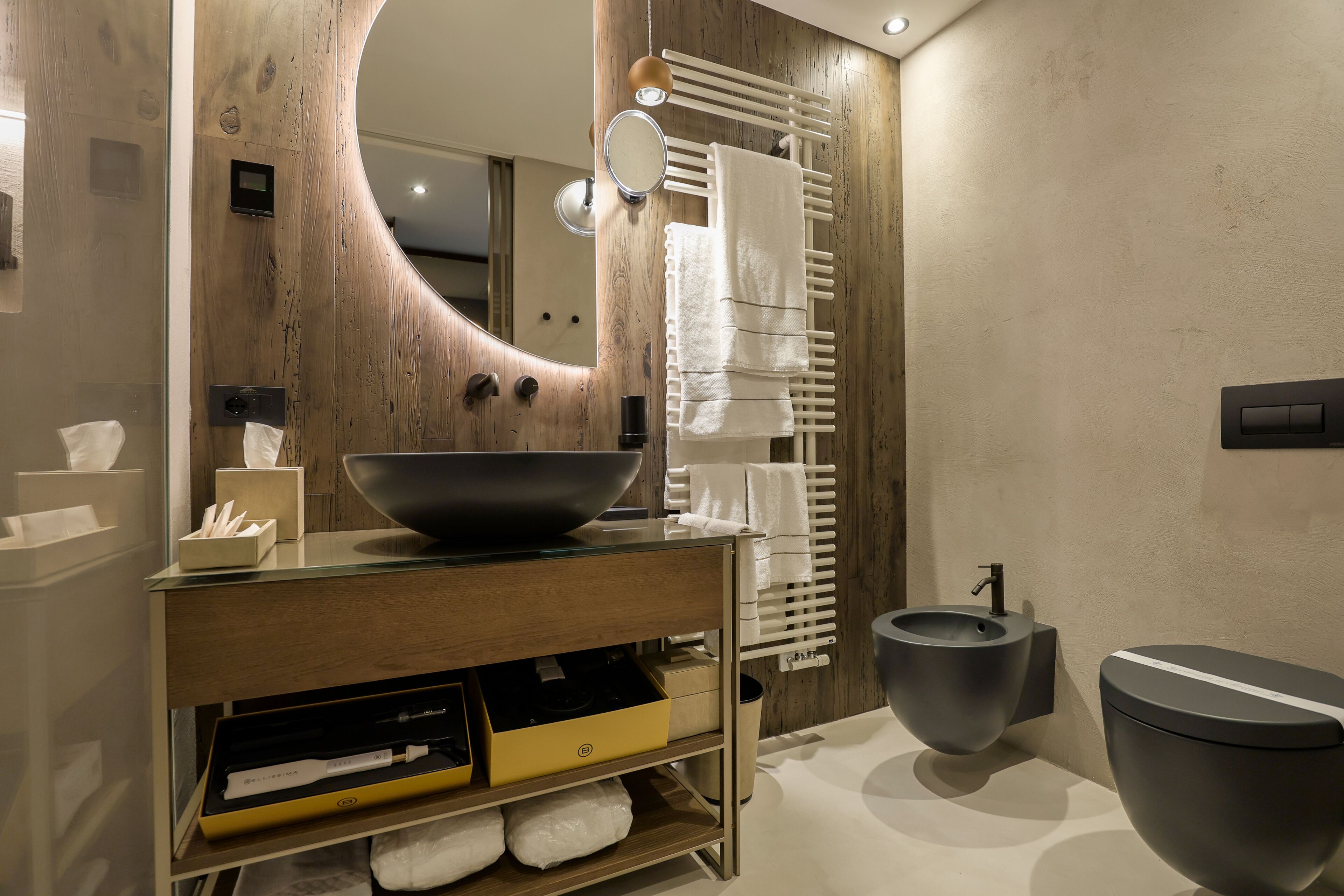Un baño moderno con un mueble de tocador de madera, un espejo grande, un lavabo negro, un inodoro y un perchero para toallas.
