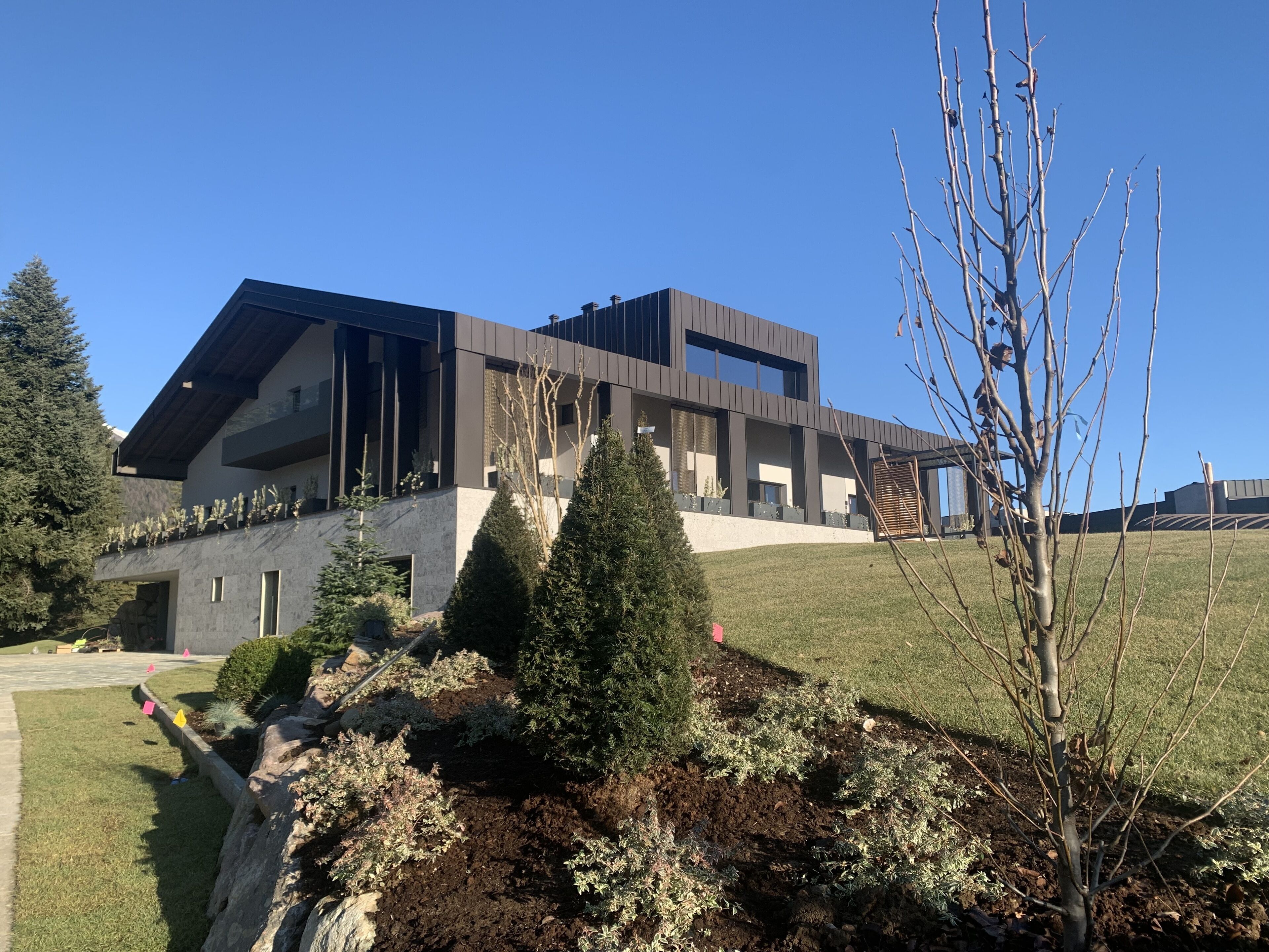 A modern building with a sloped roof, large windows, and a well-maintained garden with various shrubs and a tree.