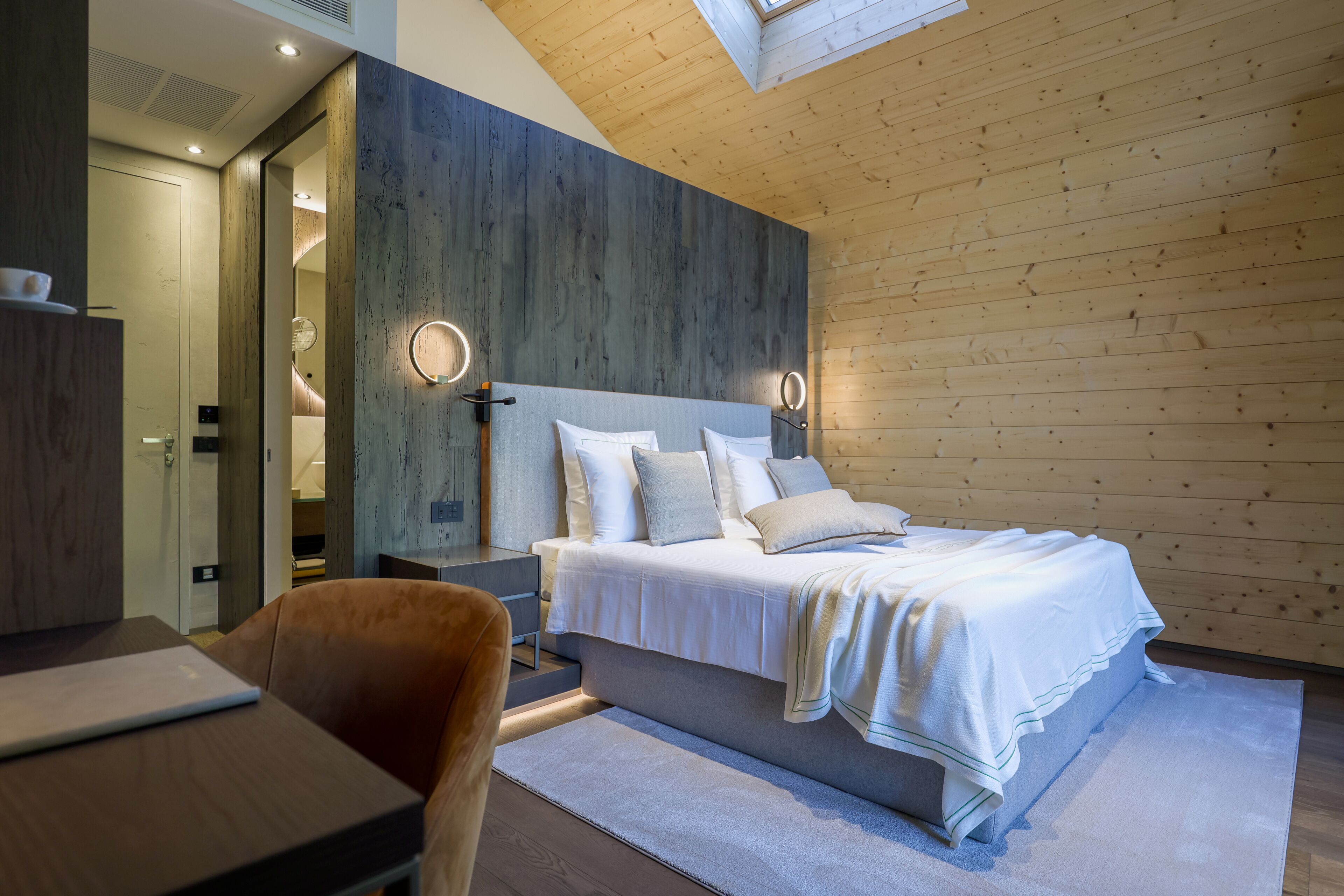 Un dormitorio moderno con techo de madera, una cama grande con ropa blanca, mesitas de noche y un escritorio con una computadora portátil.