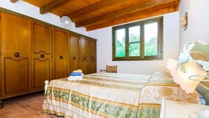 Ein Schlafzimmer mit einem großen Holzschrank, einem Bett mit floralem Überwurf, einem Fenster mit Blick auf Grünflächen und einem kleinen Tisch mit Lampe.