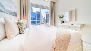 Una camera d'albergo con un letto grande, una finestra vista città, un comodino con un vaso di fiori e uno specchio sulla parete.