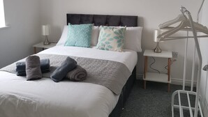 A neatly arranged bedroom with a bed, pillows, a bedside table with a lamp, a wall-mounted artwork, and a hanging rack. - Olive and Carol's Beautiful 3-bed House in Walsall (Walsall)