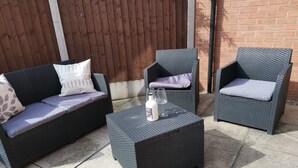 An outdoor seating area with two wicker chairs and a matching sofa, each with cushions. A bottle and glasses are placed on a square table. - Olive and Carol's Beautiful 3-bed House in Walsall (Walsall)