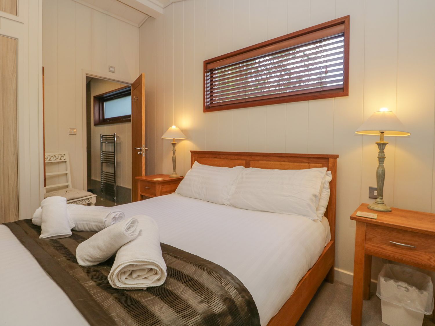 A neatly made bed with white linens and a brown striped blanket, two bedside lamps, a wooden nightstand, a wooden headboard, and a window with blinds.