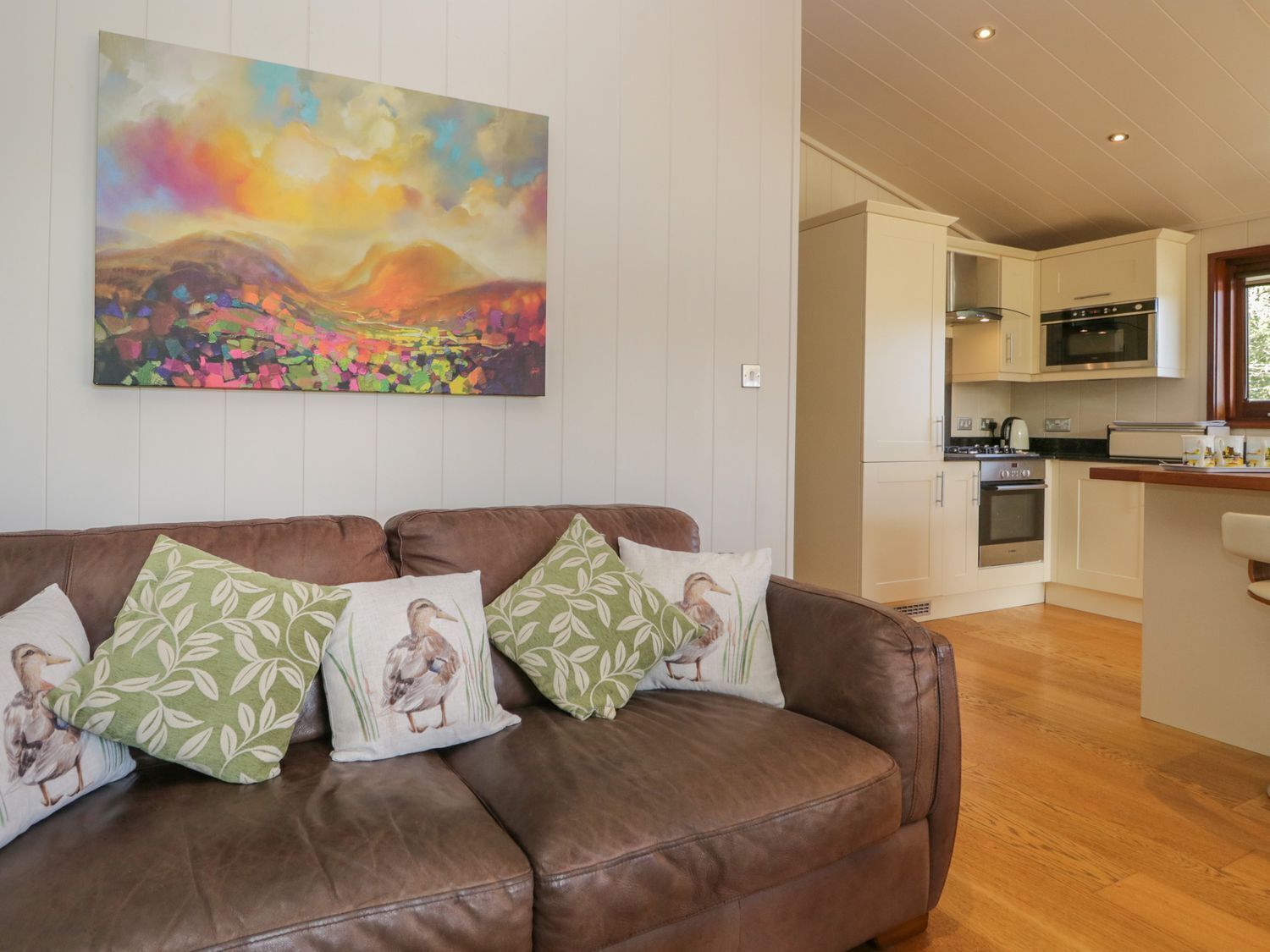 A brown leather sofa with duck-themed pillows, a colorful abstract painting, and a modern kitchen with white cabinets and a wooden countertop.