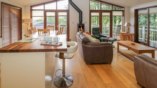 A spacious living area with a wooden floor, a fireplace, a dining table with chairs, and large windows. - Leafy Nook Lodge (Ambleside)