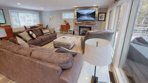 A living room with a sofa, armchair, and ottoman, a fireplace, a television, and large windows. - Lakefront Haven • Dock, Fishing & Swim (Cadiz)