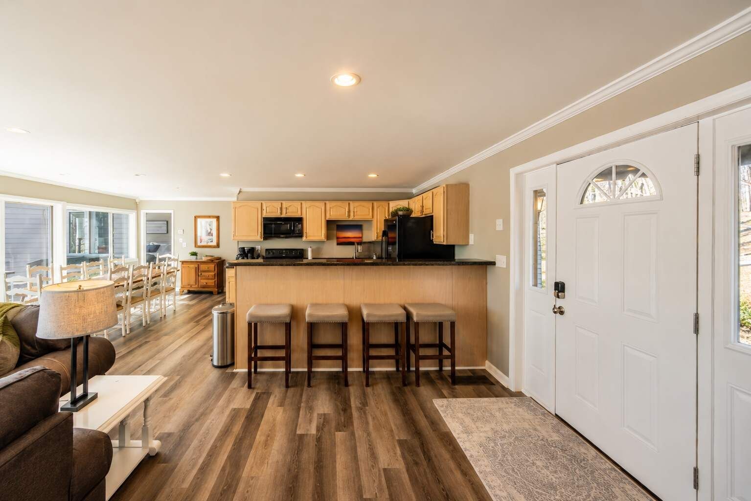 A modern kitchen with wooden cabinets, a central island with stools, and a dining area with a sofa and lamp.