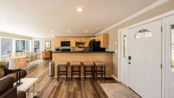 A modern kitchen with wooden cabinets, a central island with stools, and a dining area with a sofa and lamp.