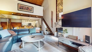 A spacious living room with a leather sofa, a round coffee table, and a TV on a wooden stand. A staircase leads to a kitchen area with white cabinets and a stone accent wall. - Pet-Friendly, Renovated, Spacious, Great Lighting. Private Gas Grill, Laundry (Mammoth Lakes)