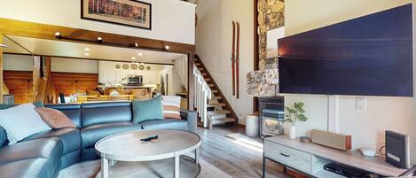Un amplio salón con un sofá de cuero, una mesa de centro redonda y un televisor sobre un mueble de madera. Una escalera conduce a una zona de cocina con armarios blancos y una pared decorada con piedra.