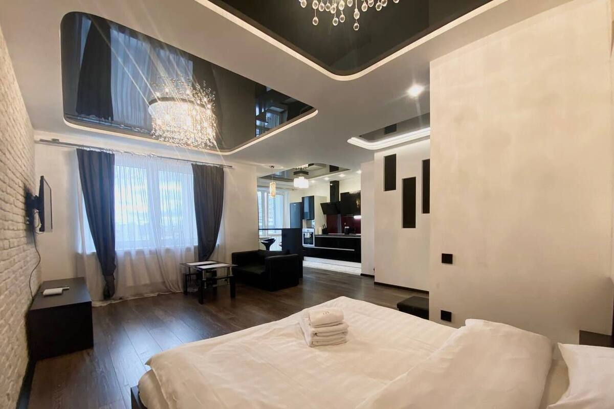 A modern hotel room with a large bed, a desk, a chair, and a view of the kitchen area.
