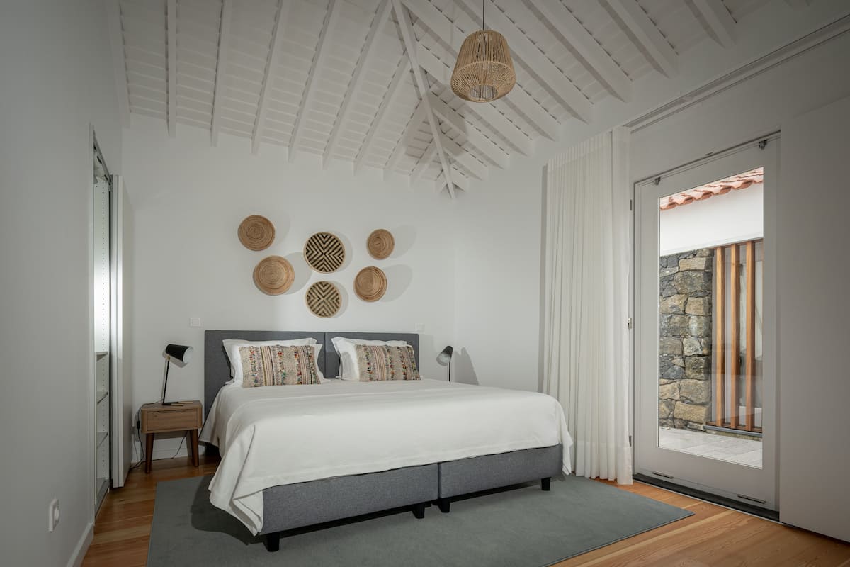 Quarto com cama, mesas de cabeceira, luminária na parede e vista através de uma porta de vidro.