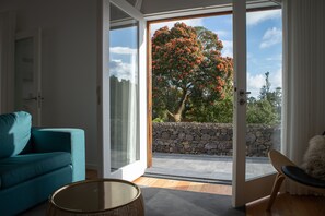 Un salon moderne avec un canapé de couleur turquoise, une table ronde en verre et une vue sur un mur de pierre et un arbre, visibles à travers une porte vitrée.