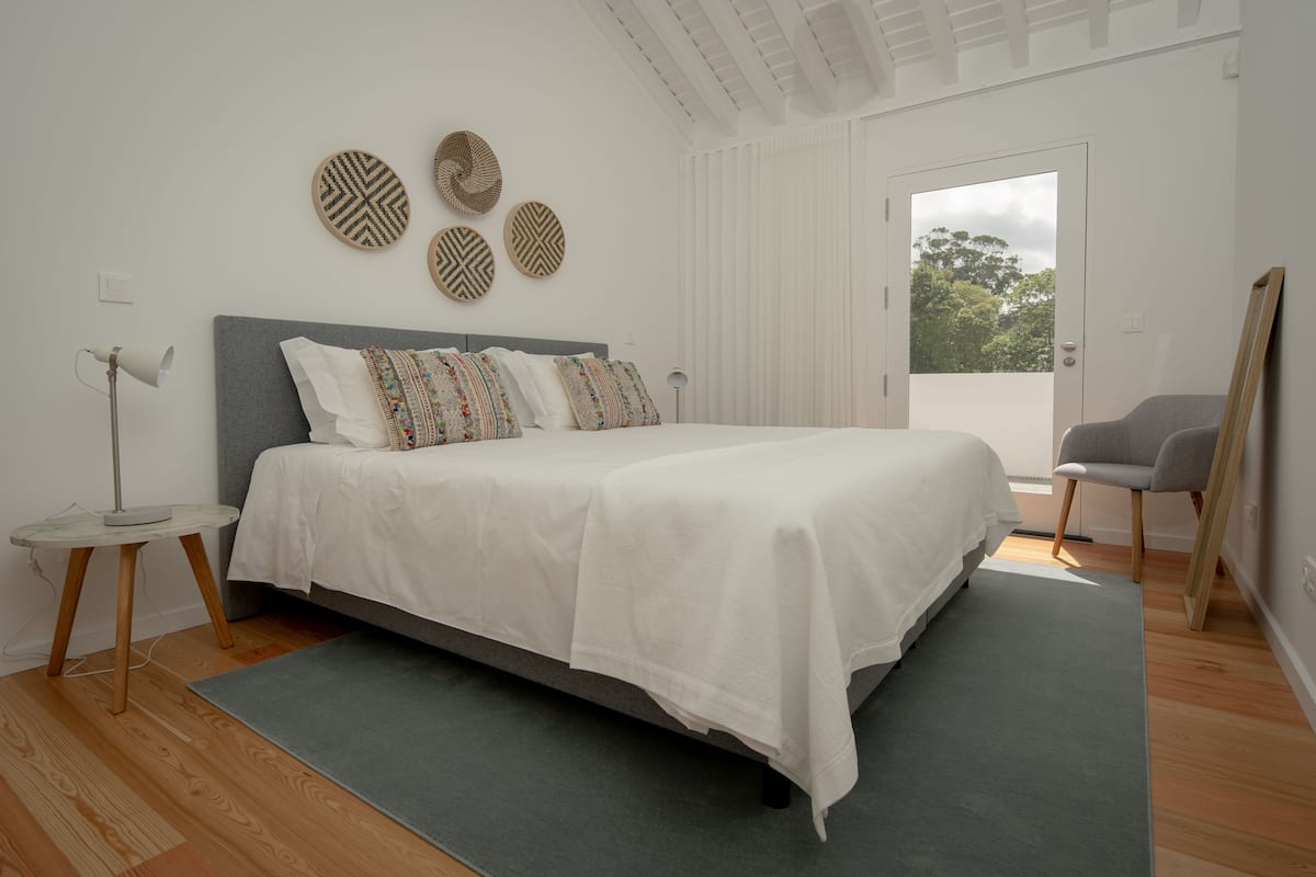 Quarto com uma cama, uma mesa redonda, uma cadeira e uma porta que dá para uma varanda.