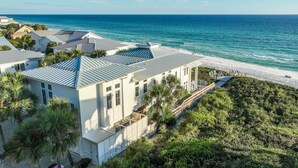 A coastal property with a large building featuring a sloped roof, surrounded by lush greenery and a beach.