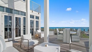 A modern outdoor deck with white furniture, a view of the ocean, and a building with large glass doors.