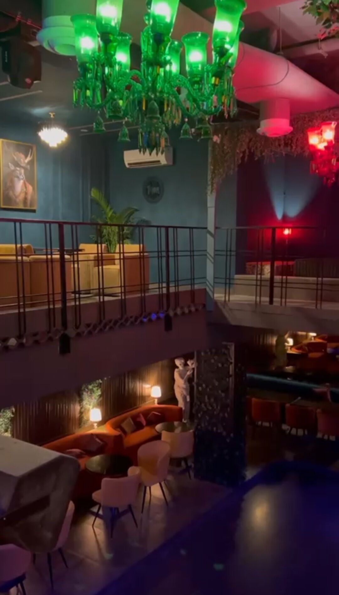 Um hall de hotel com vários níveis, com um candelabro verde, uma parede azul com uma pintura de um veado, e um nível inferior com um sofá vermelho e uma pequena mesa.