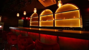 A bar area with red upholstered chairs, a counter, and three illuminated arched windows. - Hotel Thor (Agra)